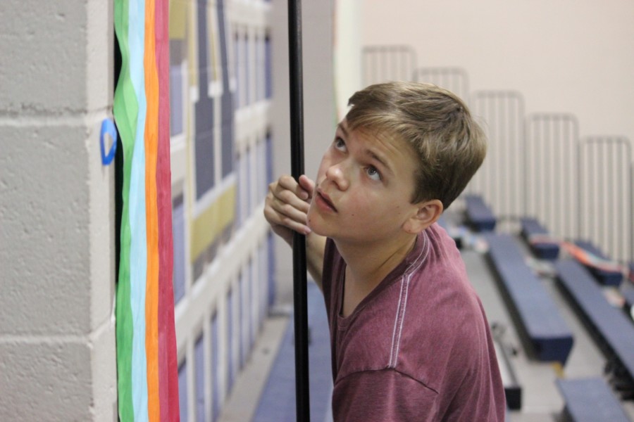 Sophomore Nathan Gray uses a paint roller to place a rainbow poster, one of the many Candyland themed decorations, on a column in the gym.