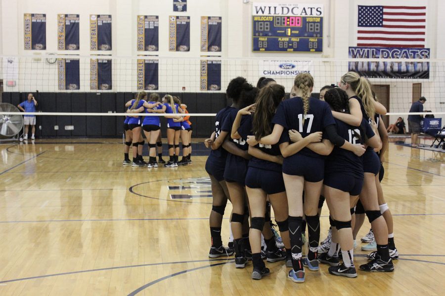 Varsity girls huddle up with their opponents behind them.